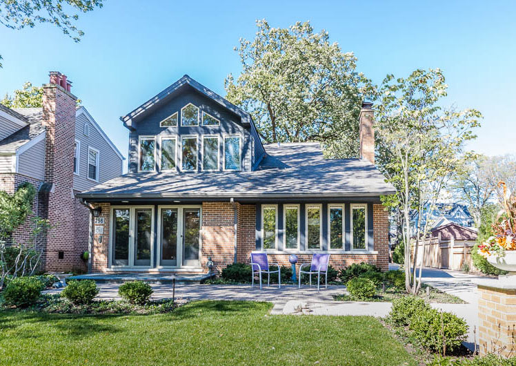 two story brick and blue home with many windows two chairs on back patio and green yard with shrubs