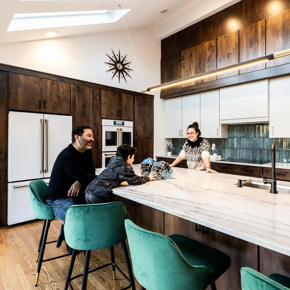 A modern luxury kitchen remodel with dark wood cabinets, white countertops, and a large island with green velvet barstools. A smiling family of three—a man, a woman, and a young boy—gather around the island, engaged in conversation. The boy has hockey gear on the counter, and a sleek pendant light hangs above the island. The kitchen features a skylight, a stylish clock on the wall, and a combination of open and closed cabinetry